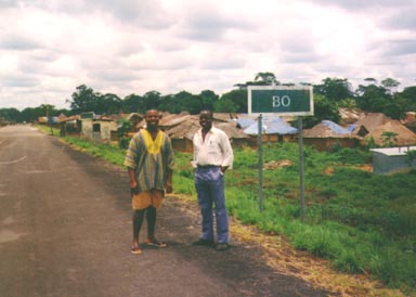 Two colleagues, Mr. Massaquoi and Boy Ru, at the road sign of our village, Bo Waterside, mere steps from the river that divides Liberia and Sierra Leone. Mr. Massaquoi was the leader of Men United to Save Humanity. Rufus and I were both mechanics; we spent many long evenings figuring-out many different kinds of problems. Note the blue UNHCR tarps covering holes in the roofs behind them. When the rainy season arrived, we needed lots more tarps. Photo by Tom Burwell.